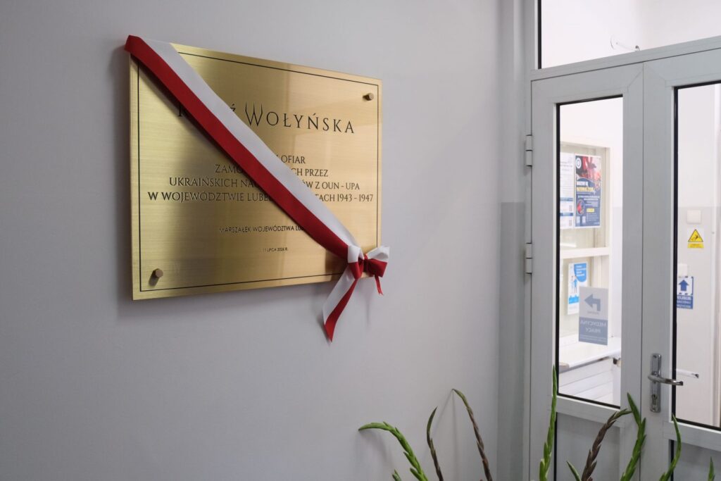 Odsłonięta tablica pamiątkowa na ścianie budynku, przewiązana czerwono-białą wstęgą