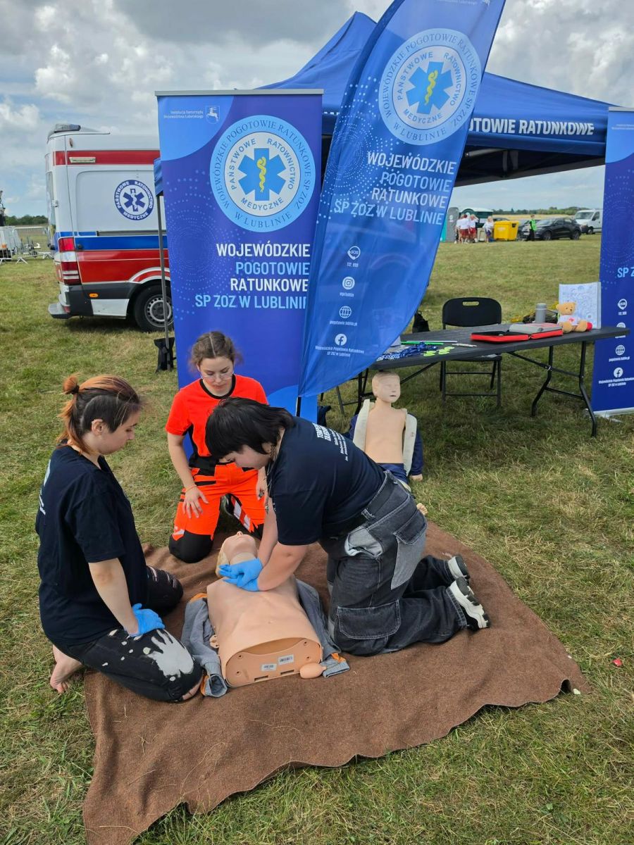 Nauka pierwszej pomocy na fantomie przy stoisku pogotowia, w tle ambulans