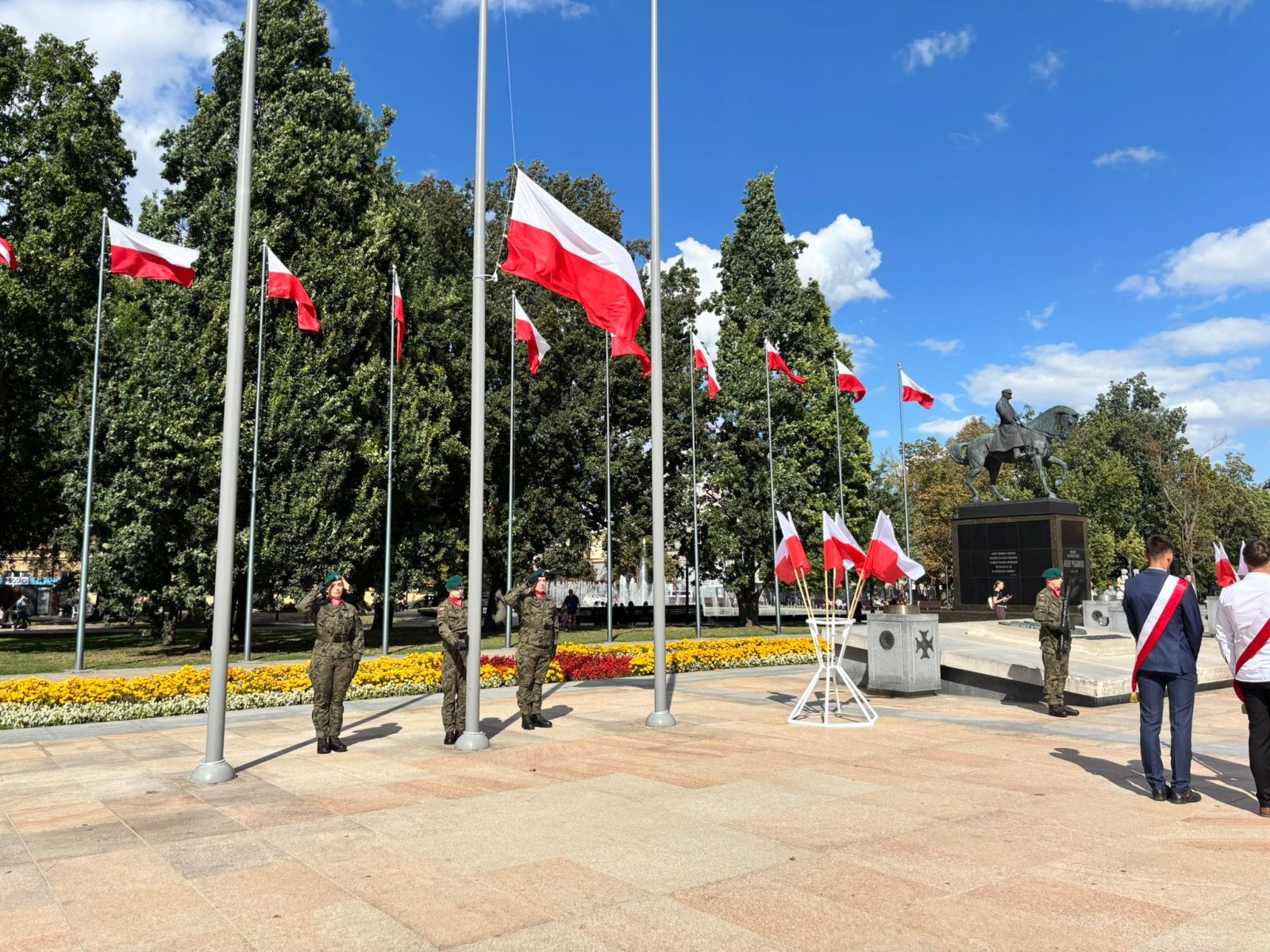 Flagi państwowe na masztach przed pomnikiem, w tle uczestnicy uroczystości i poczty sztandarowe